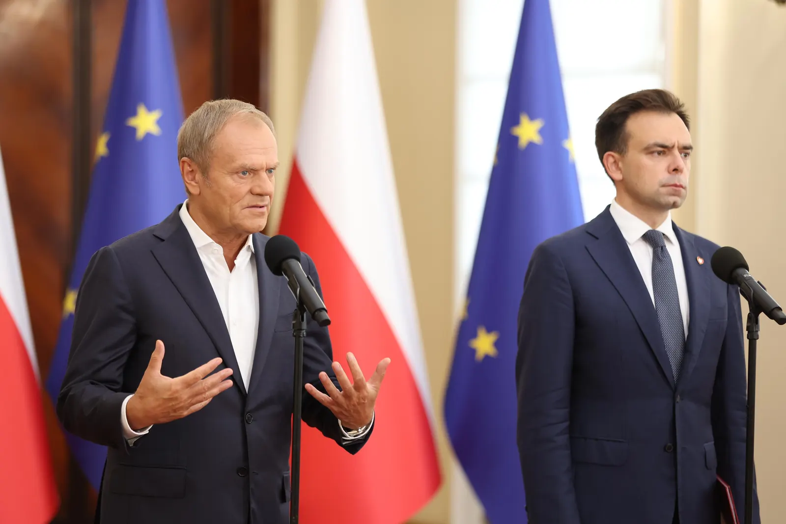 Premier Donald Tusk przemawiający podczas konferencji prasowej, z dyskretnym tłem przedstawiającym polską flagę.